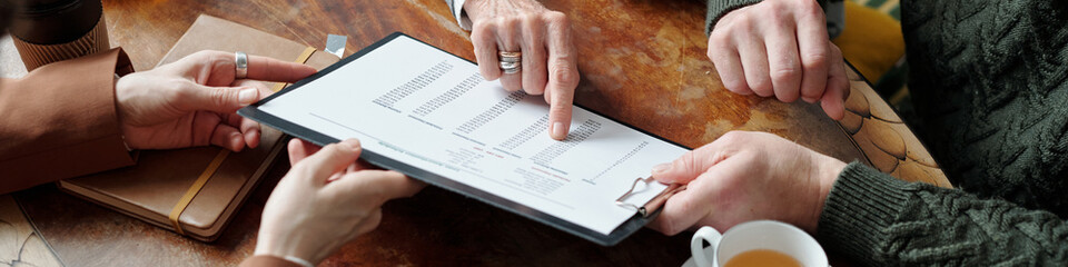 Senior hands pointing at business contract on clipboard during casual meeting. Coffee cups and notepad on wooden table enhancing relaxed atmosphere