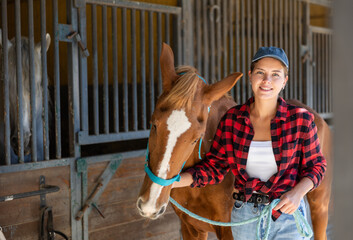 Young woman leading tethered brown horse out of stable