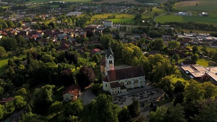 Drone panorama of a peaceful Bavarian village with a historic Catholic church and cemetery in Ortenburg. Summer sunlight, trees, and scenic countryside complete this spiritual landscape. 