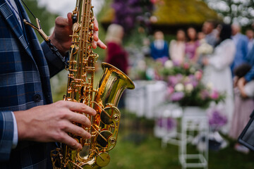 Close-up of a saxophonist playing at an outdoor wedding ceremony, with blurred guests, flowers, and...