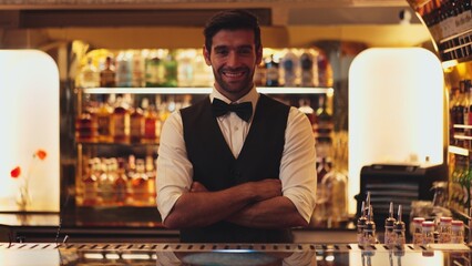 Profile good looking smiling smart bartender wiping glass at luxurious beverage counter bar scene looking at camera to pose neat professional barman costume greeting warm at bar nightclub. Vinosity.