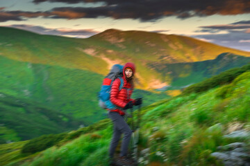 Motion Blur of Female hiker in red jacket and hood standing on windy mountain slope at golden hour. Thoughtful gaze, trekking poles, dramatic light over green Carpathian hills in background