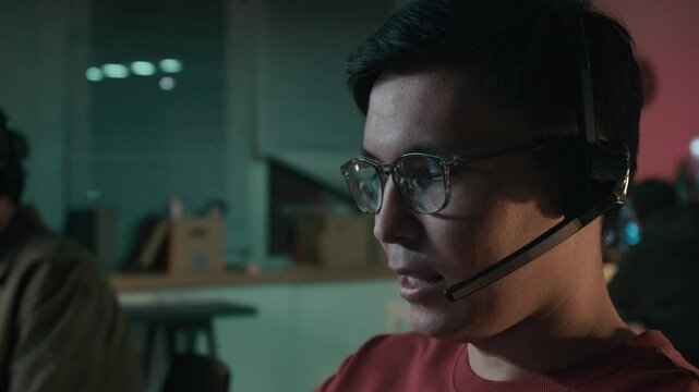 Close-up of face of grinning young Asian male scammer in glasses working at computer at illegal call center, impersonating officer, talking confidently into headset to target person to reveal password