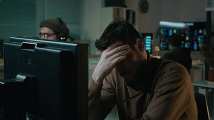 Medium shot of dismayed Caucasian scam caller sitting in illegal call center, staring at computer screen after failing to swindle victim with fake investment, feeling tired and rubbing face