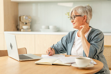 Middle aged senior woman using laptop computer writing notes at home. Focused mature old businesswoman work in home office making written records doing online work. Mature boss professional worker