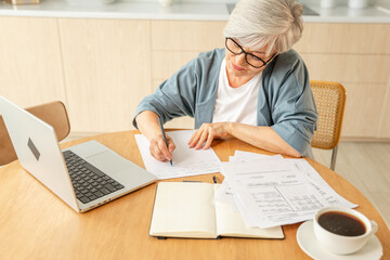 Middle aged senior woman using laptop computer writing notes at home. Focused mature old businesswoman work in home office making written records doing online work. Mature boss professional worker