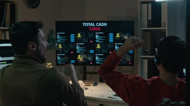 Medium rear shot of two male scam call center supervisors sitting on chairs in front of large screen, watching money being transferred into criminals bank account, cheering with joy and bumping fists