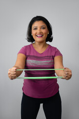 Happy fitness woman exercising arms with a green rubber band. Ideal for home workout, muscle strengthening and injury recovery. Studio portrait, neutral gray background with copy space.