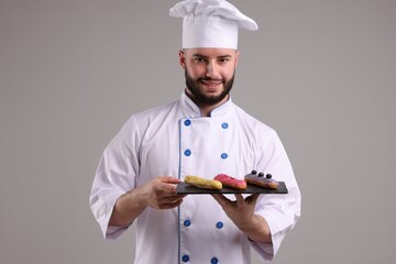 Happy confectioner in uniform holding delicious eclairs on light grey background