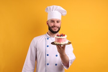 Happy confectioner in uniform holding cake with berries on orange background