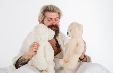 Man with two teddy bears. Bearded man playing in bed with teddy bears. Smiling hipster in bathrobe...