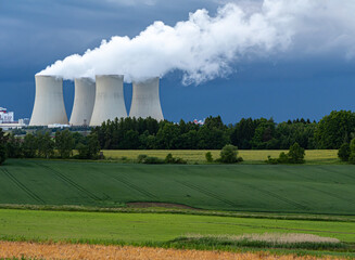 Nuclear power plant cooling towers emitting steam above lush green fields and forest, under a...