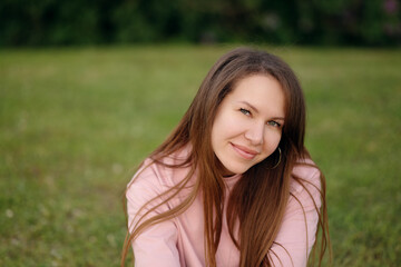 Close-up of a smiling young woman sitting on green grass in a spring park. Natural look, long hair, hoop earrings, and pink T-shirt. Cheerful and relaxed outdoor portrait with soft light.