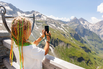 Back view little cute adorable blond kid girl braids hair take photo scenic Grossglockner high mountain alpine road landscape Austria sunny summer day. Kid shooting nature with smartphone