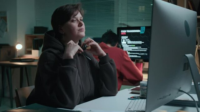 Medium shot of scruffy young Caucasian female scam call center employee talking politely to target on internet phone, finishing conversation, taking off headset, rubbing face, tired after night work