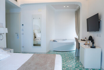 Bright hotel room in Amalfi, Italy, featuring a large bed, green and white tiled floor, wall mounted TV, desk with coffee machine, and a white bathtub.