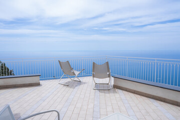 A spacious terrace with tiled flooring and white railing overlooks the Tyrrhenian Sea. Two minimalist lounge chairs face the water under a partly cloudy sky.