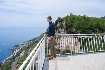 A man stands on a tiled terrace with a white railing, overlooking the Amalfi Coast. The view includes the Tyrrhenian Sea, cliffs, and coastal buildings.