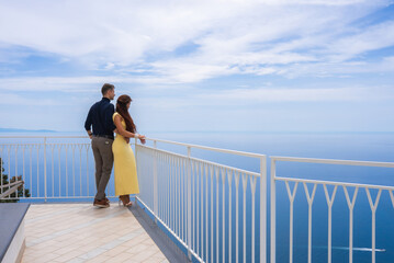 A couple stands on a tiled terrace with a white railing, gazing at the Tyrrhenian Sea under a...