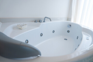 A sleek white jacuzzi tub with jet nozzles, chrome faucet, and headrest. Toiletries in a basket sit on the edge, with soft light through a sheer curtain.