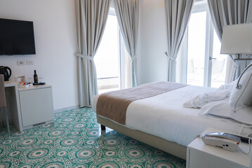Bright hotel room with white linens, tiled floor, desk area, and glass doors opening to a balcony....