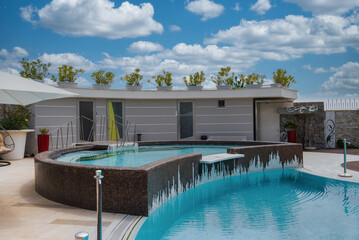 Luxurious outdoor pool area with cascading water, tiled deck, contemporary furnishings, and a sleek building under a bright blue sky in Amalfi, Italy.