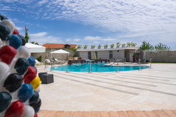 Outdoor pool with tiled deck, white umbrellas, lounge chairs, and a colorful sculpture. Modern building, stone wall, and greenery in the background.
