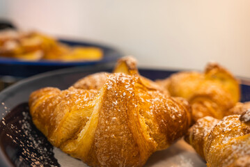 Golden croissants dusted with powdered sugar sit on a dark plate, with blurred pastries in the background creating a warm dining atmosphere.