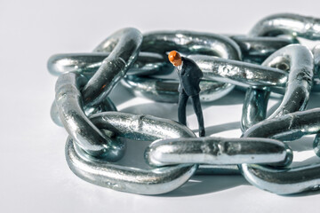 A miniature businessman figurine stands amidst the links of a large metal chain on a white surface. The scene suggests a concept related to business, security, or obstacles.