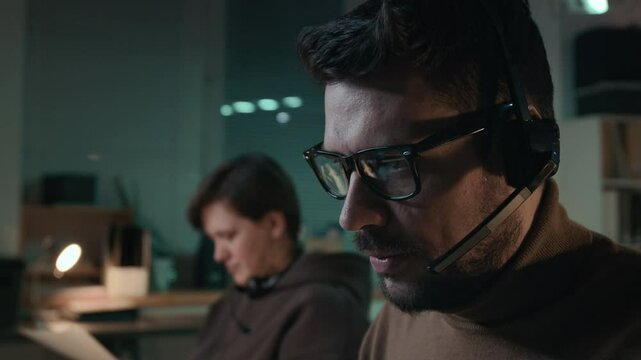 Side close-up of young Caucasian male scammer wearing glasses, headset sitting at desk in criminal call center, talking on phone to victim while impersonating government official to extract passwords