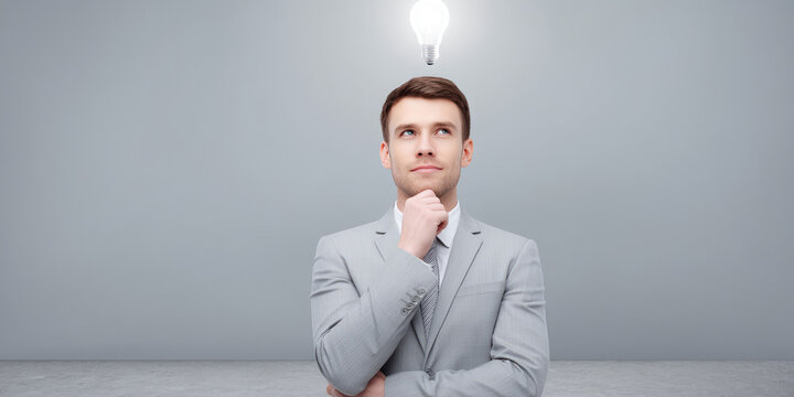 Thoughtful man in gray suit with hand on chin gazes upward at lit bulb above head, symbolizing a bright idea or inspiration. 
