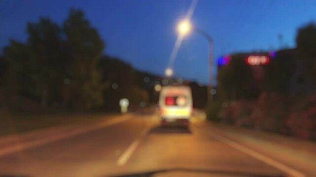 Blurred night video of a speeding ambulance with flashing lights moving through traffic, captured from behind in a dramatic pursuit view