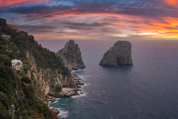 Faraglioni rocks rise from the sea near Capri, Italy, with cliffs lined by greenery and whitewashed buildings under a vibrant orange and pink sunset. © Aerial Film Studio