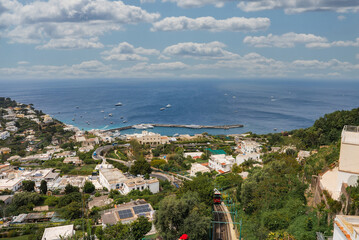 View of Capri, Italy featuring a harbor with boats, whitewashed buildings, lush greenery, a winding road, and a funicular railway under a partly cloudy sky. © Aerial Film Studio