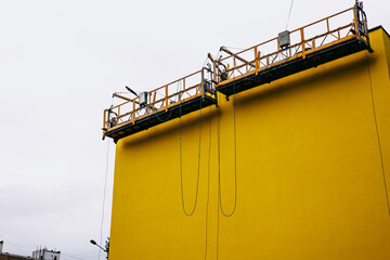 Scaffolding against a vibrant yellow wall with ropes hanging down against an overcast sky. This...