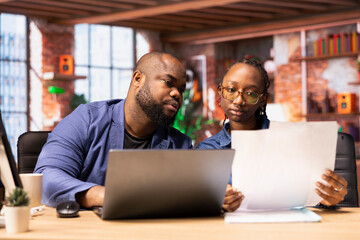 Young people completing spreadsheets on laptop by entering numerical data from papers, calculating totals and analyzing figures. African american couple working together from home.