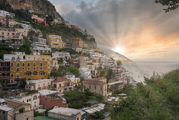 Obraz premium Colorful buildings cascade down a hillside in Positano, Italy, with a sunset illuminating the sky, lush greenery, and boats on calm coastal waters.