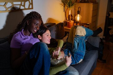 Young interracial lesbian couple enjoying a movie night at home, eating popcorn from a bowl while relaxing on their comfortable sofa