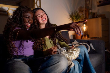 Two young women are sitting on a sofa, enjoying a movie night with popcorn, sharing laughter and pointing at the screen