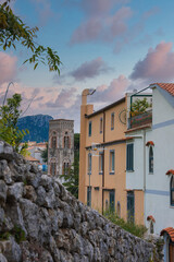A stone bell tower with arched windows stands in Ravello, Italy, surrounded by pastel buildings, terracotta roofs, greenery, and a pink blue sky.