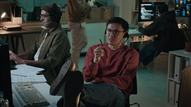 Medium full shot of young male fraudster relaxing on chair in front of computer, thinking of effective approach to extort money from victim, rehearsing dialogue lines before making scam phone call