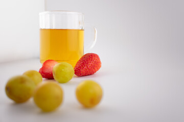 Clear glass mug with golden herbal tea next to a wooden coaster holding fresh strawberries and green grapes. The arrangement suggests a focus on health and nutrition, with elegant presentation.