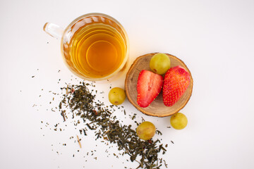 Clear glass mug with golden herbal tea next to a wooden coaster holding fresh strawberries and green grapes. The arrangement suggests a focus on health and nutrition, with elegant presentation.