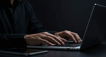 A person in a black shirt typing on a laptop keyboard in a dark room with a tablet nearby on the table