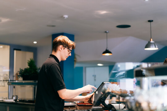 Young waiter serving customer at cash point in cafe. Man working with POS terminal. Student working part-time. Problems of unemployment and Job opportunities for youth. Small business vacancies