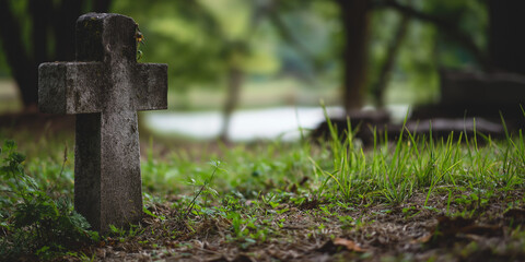 Weathered stone cross grave in forested cemetery with green grass and blurred lake in background
