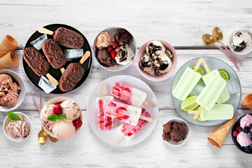 Cool summer food table scene. Variety of ice cream bowls, popsicles and frozen treats. Above view on a white wood background.