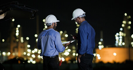 Two college Industrial engineer with white safety hat work with digital tablet and walkie talkie on night shift at Refinery and gas petrochemical plant, Industrial and business