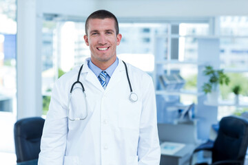 Male physician smiling while standing in exam room by desk with computer monitor and stethoscope