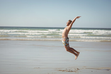 Man jumping above wet sand near shoreline at low tide, wearing colorful floral swim trunks